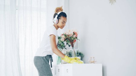 Photo for Cheerful maid African American girl is doing housework dusting using cloth and wearing protective gloves, young woman is listening to music in headphones - Royalty Free Image