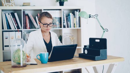 Téléchargez les photos : Jolie jeune femme travaille dans un bureau moderne assis à table, en utilisant un ordinateur portable et en écrivant dans un cahier. Des étagères avec papiers, plantes vertes et livres sont visibles - en image libre de droit