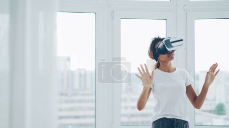 Téléchargez les photos : Fille afro-américaine en vêtements décontractés utilise des lunettes de réalité virtuelle et des gestes tout en se tenant dans son appartement avec des fenêtres en arrière-plan. Concept moderne de technologie, de divertissement et de personnes
. - en image libre de droit