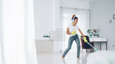 Photo for Pretty African American girl cheerful maid is cleaning floor in beautiful flat with plastic mop and listening to music, singing and dancing. Routine, house and fun concept. - Royalty Free Image