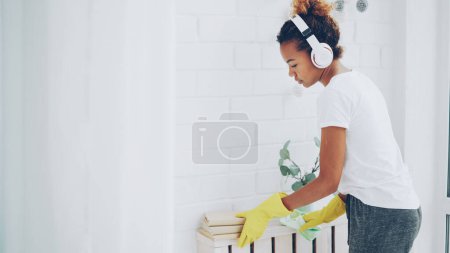 Foto de Alegre criada afroamericana chica está haciendo tareas domésticas desempolvar usando tela y usar guantes de protección, mujer joven está escuchando música en los auriculares - Imagen libre de derechos