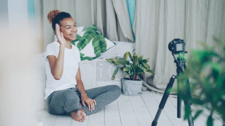 Téléchargez les photos : Jolie adolescente afro-américaine vlogger enregistre une vidéo pour les adeptes des médias sociaux, la fille regarde la caméra sur le trépied, parlant et gesticulant assis sur le sol de la chambre
. - en image libre de droit