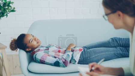 Téléchargez les photos : Psychologue attentif est en consultation avec la jeune femme afro-américaine, écouter et prendre des notes alors que le client est couché sur le canapé et parler avec le sourire
. - en image libre de droit