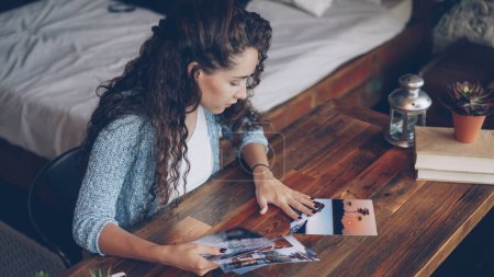 Photo for Pretty woman female photographer is looking at photographs sitting at table and putting pictures on wooden desk. Loft style apartment with modern furniture is visible. - Royalty Free Image