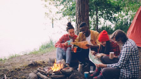 Photo for Happy young people friends are looking at maps sitting in forest around fire and holding drinks. Flame is moving, tent and trees are visible. - Royalty Free Image
