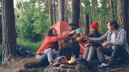 Téléchargez les photos : Touristes insouciants sont griller, tasses et verres clinquant avec des boissons chaudes puis boire assis autour du feu dans la forêt près de la tente. Concept d'amitié, de loisirs et de jeunesse
. - en image libre de droit