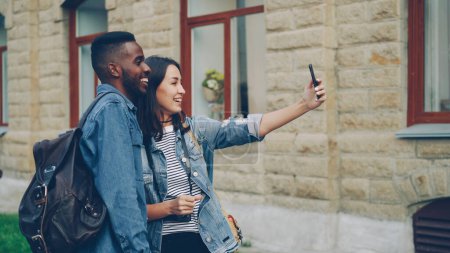 Foto de Emocionado turistas joven hombre y mujer están haciendo videollamada en línea con dispositivo de retención de teléfonos inteligentes y hablando mostrando pulgares hacia arriba expresando emociones positivas. - Imagen libre de derechos