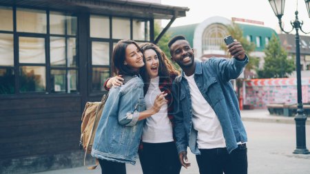 Photo for Happy African American guy is taking selfie with friends beautiful Caucasian girls standing in street posing and holding modern smartphone during journey in foreign country. - Royalty Free Image