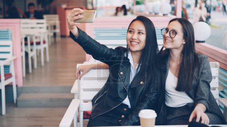 Foto de Hermosas mujeres jóvenes están tomando selfie en la cafetería sentado en la mesa y el uso de teléfono inteligente. Atractiva chica caucásica está usando gafas modernas, su amigo asiático está sosteniendo gadget
. - Imagen libre de derechos