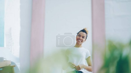 Photo for Pretty African American girl looking in the mirror at home and smiling. Beautiful young people, youth lifestyle and interiors concept. - Royalty Free Image