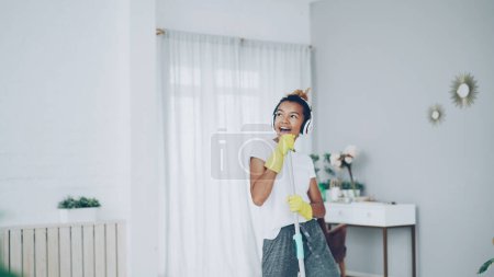 Téléchargez les photos : Funny fille afro-américaine apprécie la musique dans les écouteurs et faire des travaux ménagers plancher de lavage à la maison, jeune femme danse et chante en s'amusant. Les gens et le concept technologique
. - en image libre de droit
