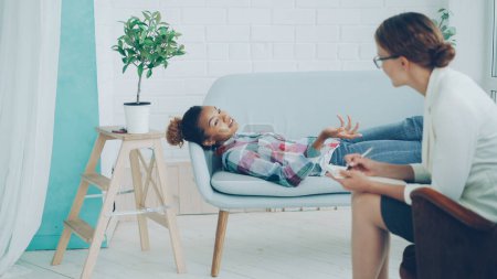 Téléchargez les photos : Un adolescent afro-américain détendu parle à un psychologue allongé sur un canapé et souriant pendant que le thérapeute écoute et prend des notes. Jeunes, santé mentale et concept de profession
. - en image libre de droit