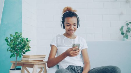 Foto de Mujer afroamericana feliz está navegando por Internet con el teléfono inteligente y riendo sentado en cómodo sofá en casa, chica está escuchando música a través de auriculares
. - Imagen libre de derechos