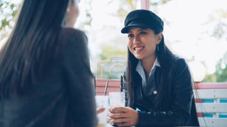 Photo for Happy Asian girl is talking to her female friend in street cafe then toasting and clinking glasses with cocktails enjoying meeting. Communication, friendship and drinks concept. - Royalty Free Image