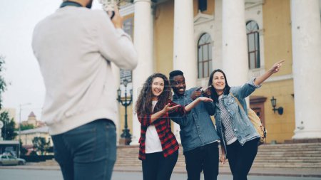 Photo for Professional photographer is shooting multiracial group of travelers happy young people posing for camera in front of beautiful theater with backpacks and laughing. - Royalty Free Image
