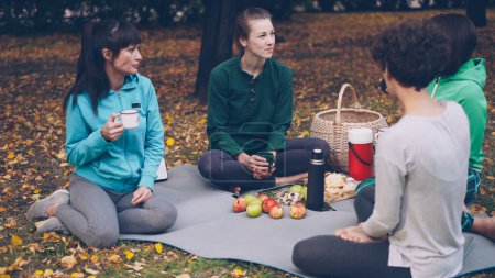 Foto de Hermosa sportswomen son relajantes después de entrenar al aire libre teniendo picnic en esteras comer bocadillos y beber té. Las niñas están hablando y riendo disfrutando de la naturaleza otoño
. - Imagen libre de derechos