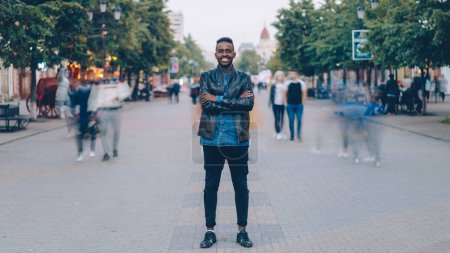 Photo for Portrait of happy African American guy wearing stylish jeans and leather jacket standing alone in street downtown, smiling and looking at camera with crowd moving by. - Royalty Free Image