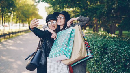 Photo for Excited female friends are taking selfie using smartphone and holding shopping bags with purchases on sunny autumn day. Modern technology and youth lifestyle concept. - Royalty Free Image
