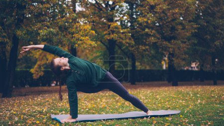 Foto de La joven esbelta está haciendo yoga al aire libre cambiando asanas y disfrutando del movimiento, el aire fresco y la hermosa naturaleza otoñal. Millennials, estilo de vida saludable y concepto de recreación
. - Imagen libre de derechos