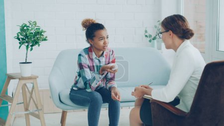 Photo for Emotional African American lady is talking to psychologist sitting on couch, talking and gesturing during professional consultation while specialist is listening holding paper and pen. - Royalty Free Image
