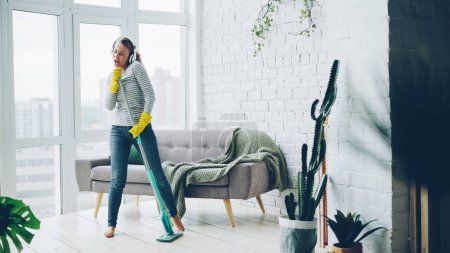 Photo for Happy young housewife is dancing and singing during housecleaning, she is listening to music in headphones and mopping floor having fun. Housework and technology concept. - Royalty Free Image