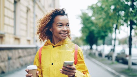 Photo for Happy African American woman is using smartphone touching screen and smiling walking outdoors in beautiful city with to-go coffee. Modern lifestyle, people and communication concept. - Royalty Free Image