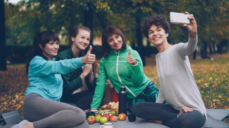 Foto de Chicas atractivas yogini están tomando selfie usando teléfono inteligente durante el picnic en el parque en otoño. Las niñas están posando y sonriendo divirtiéndose sentados juntos en esteras
. - Imagen libre de derechos