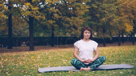 Photo for Pretty young woman is relaxing sitting in lotus pose on yoga mat on grass in park and breathing resting after individual practice. Meditation and nature concept. - Royalty Free Image