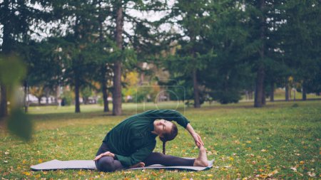 Photo for Smiling young woman is exercising in park doing yoga sitting on mat on green and yellow grass enjoying fresh air and healthy activity. Youth, active lifestyle and recreation concept. - Royalty Free Image