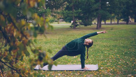 Foto de Estudiante de yoga femenino está practicando variaciones de la posición de ángulo lateral Parsvakonasana en la esterilla de yoga en el parque. Árboles verdes y amarillos, hojas y hierba son visibles
. - Imagen libre de derechos