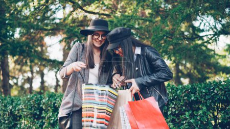 Foto de Dos mujeres jóvenes adictas a las compras están hablando en la calle en la ciudad mirando las compras en bolsas y expresando emoción. Compras, personas y concepto de felicidad
. - Imagen libre de derechos