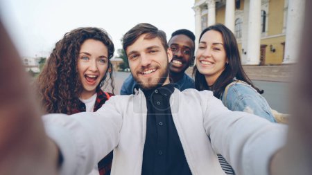 Foto de Punto de vista plano del grupo multirracial de turistas tomando selfie en el centro de la ciudad sosteniendo la cámara gadget y posando junto con gestos de la mano que expresan emociones positivas. - Imagen libre de derechos