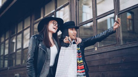 Photo for Cheerful young ladies best friends are taking selfie posing with bright paper shopping bags outdoors in the street. Young women are wearing fashionable clothing and hats. - Royalty Free Image