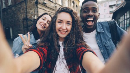 Téléchargez les photos : Point de vue prise de vue de filles et de gars joyeux groupe multiethnique prenant selfie tenant la caméra et posant à l'extérieur pendant des vacances agréables dans la belle ville. Tourisme et photographie concept. - en image libre de droit