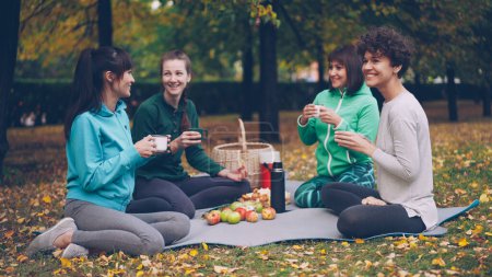 Foto de Chicas felices están teniendo picnic en el parque sentado en alfombras de yoga y comer después de la práctica al aire libre en otoño, las niñas están hablando y riendo. Comunicación, personas y concepto alimentario
. - Imagen libre de derechos