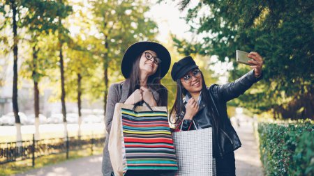 Téléchargez les photos : Des jeunes amies heureuses prennent du selfie avec des sacs en papier colorés debout à l'extérieur sur le trottoir et posant. Les filles utilisent un smartphone et portent des vêtements décontractés
. - en image libre de droit