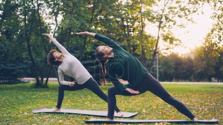 Foto de Dos mujeres guapas están haciendo yoga al aire libre en el parque de la ciudad en esteras practicando asanas y respirando aire fresco. Práctica individual, instructor profesional y concepto de naturaleza
. - Imagen libre de derechos