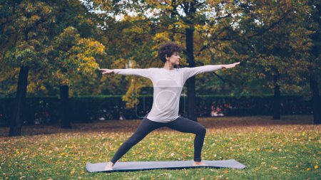 Photo for Beautiful girl with short curly hair is doing yoga outdoors standing in Warrior pose then stretching body and legs during individual practice in park. Trees and grass are visible. - Royalty Free Image