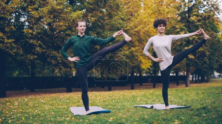 Photo for Slim young ladies are exercising outdoors in park standing on one leg practising Padangusthasana during individual class with experienced teacher. Nature and youth concept. - Royalty Free Image