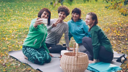 Foto de Mujeres jóvenes y bonitas se están tomando selfie con smartphone durante el picnic en el parque sentado en la hierba, posando y riendo divirtiéndose. Naturaleza, amistad y concepto tecnológico moderno
. - Imagen libre de derechos