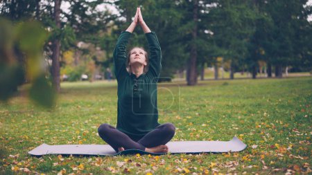 Photo for Smiling young woman is exercising in park doing yoga sitting on mat on green and yellow grass enjoying fresh air and healthy activity. Youth, active lifestyle and recreation concept. - Royalty Free Image