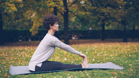 Téléchargez les photos : Slim jeune femme fait une séquence de yoga asanas assis sur le tapis dans le parc seul par une chaude journée d'automne avec de belles herbes et des arbres autour d'elle. Concept de loisirs et de sport
. - en image libre de droit