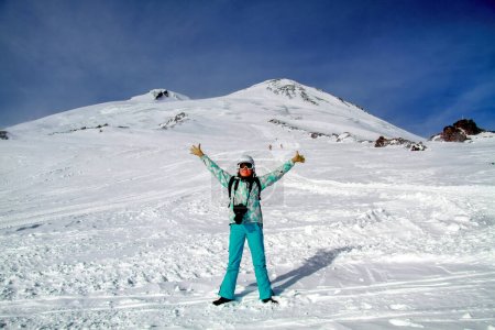 Girl skier at the mountain Elbrus. Smiling happy woman standing in a snowy mountain landscape in Caucasus stretching her arms. In background two summit of Elbrus. Adventure winter extreme sport
