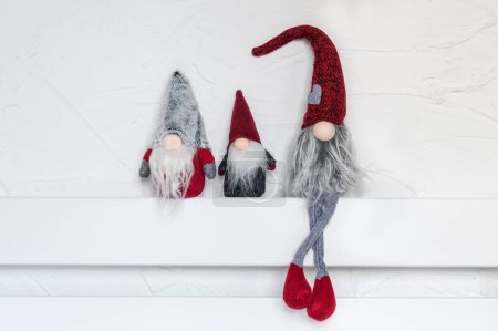 Téléchargez les photos : Elfes, décorations de Noël. Trois gnomes sont assis sur une étagère en bois blanc contre le mur. Noel et Nouvel An concept, carte de voeux, espace de copie - en image libre de droit