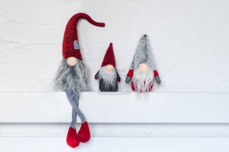Elves, Christmas decorations. Three gnomes sit on a white wooden shelf against the wall. Christmas and New Year concept, greeting card, copy space