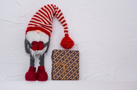 Téléchargez les photos : Gnome de Noël dans un chapeau rayé avec un cœur dans ses mains. Décoration pour votre Noël. Cadeaux sur une étagère blanche en bois contre le mur - en image libre de droit