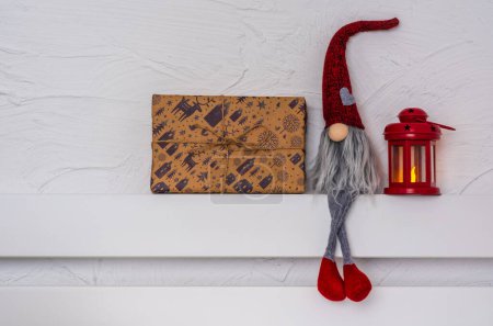Foto de Gnom de Navidad - Gonk con barba larga y sombrero rojo sentado en el estante de madera blanca con patas colgantes. Concepto de Navidad y Año Nuevo, tarjeta de felicitación, espacio para copiar - Imagen libre de derechos