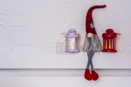 Téléchargez les photos : Noël Gnom Gonk avec longue barbe et chapeau rouge assis sur l'étagère en bois blanc avec des jambes suspendues. Noel et Nouvel An concept, carte de voeux, espace de copie - en image libre de droit