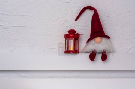 Foto de Gnom de Navidad - Gonk con barba larga y sombrero rojo sentado en el estante de madera blanca con patas colgantes. Concepto de Navidad y Año Nuevo, tarjeta de felicitación, espacio para copiar - Imagen libre de derechos