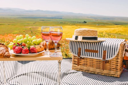 Foto de Picnic de primavera o verano con croissants, frutas y vino de rosas en el prado lleno de flores silvestres brillantes. - Imagen libre de derechos